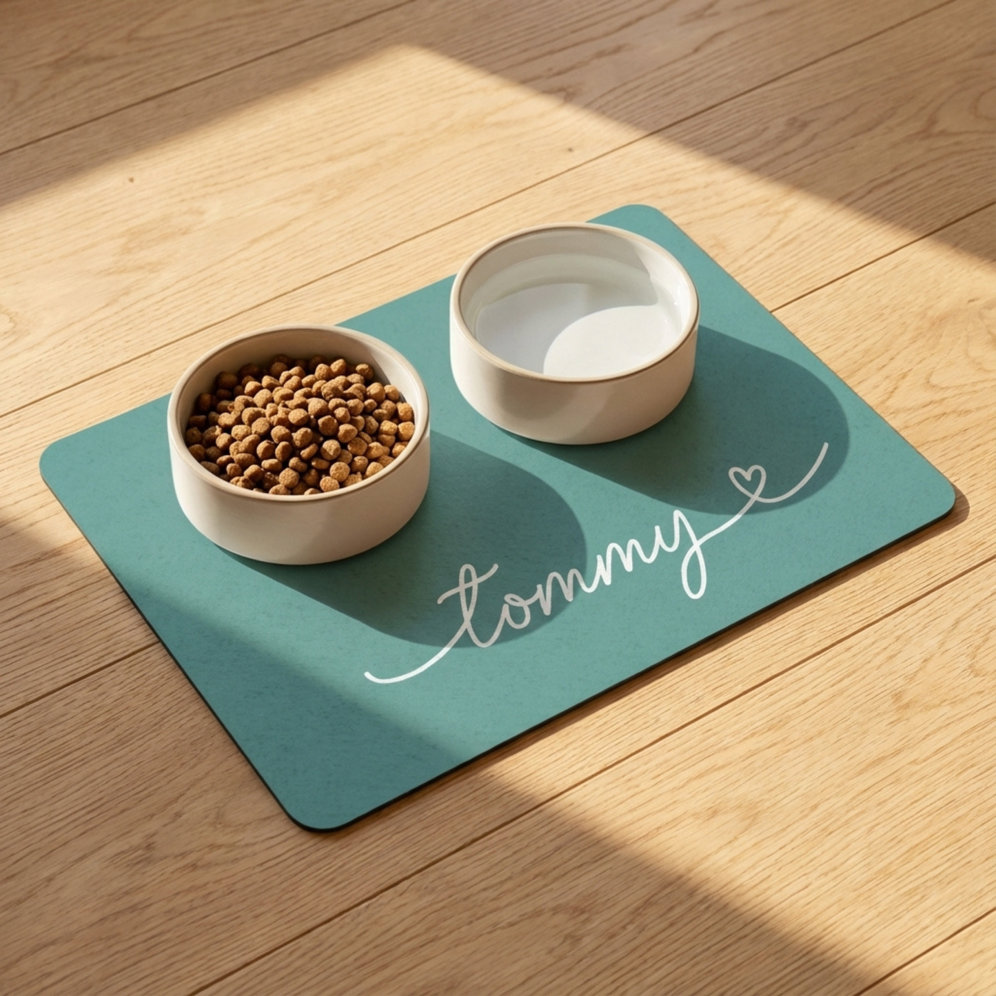 Two pet bowls on a teal mat with 'Tommy' written on it, placed on a wooden floor.
