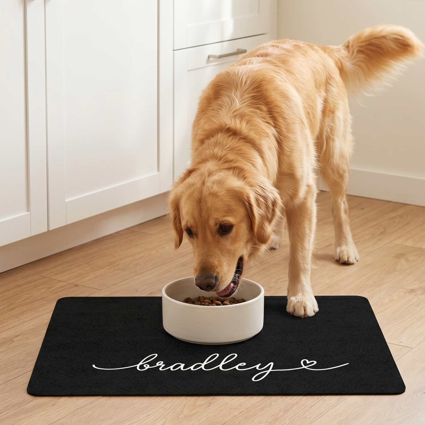 Dog eating from a bowl on a black dog mat with 'bradley' written on it in a kitchen.