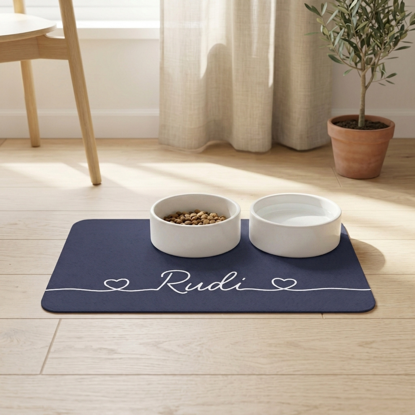 Two white pet bowls on a blue dog food mat with 'Rudi' printed on it, placed on a wooden floor.