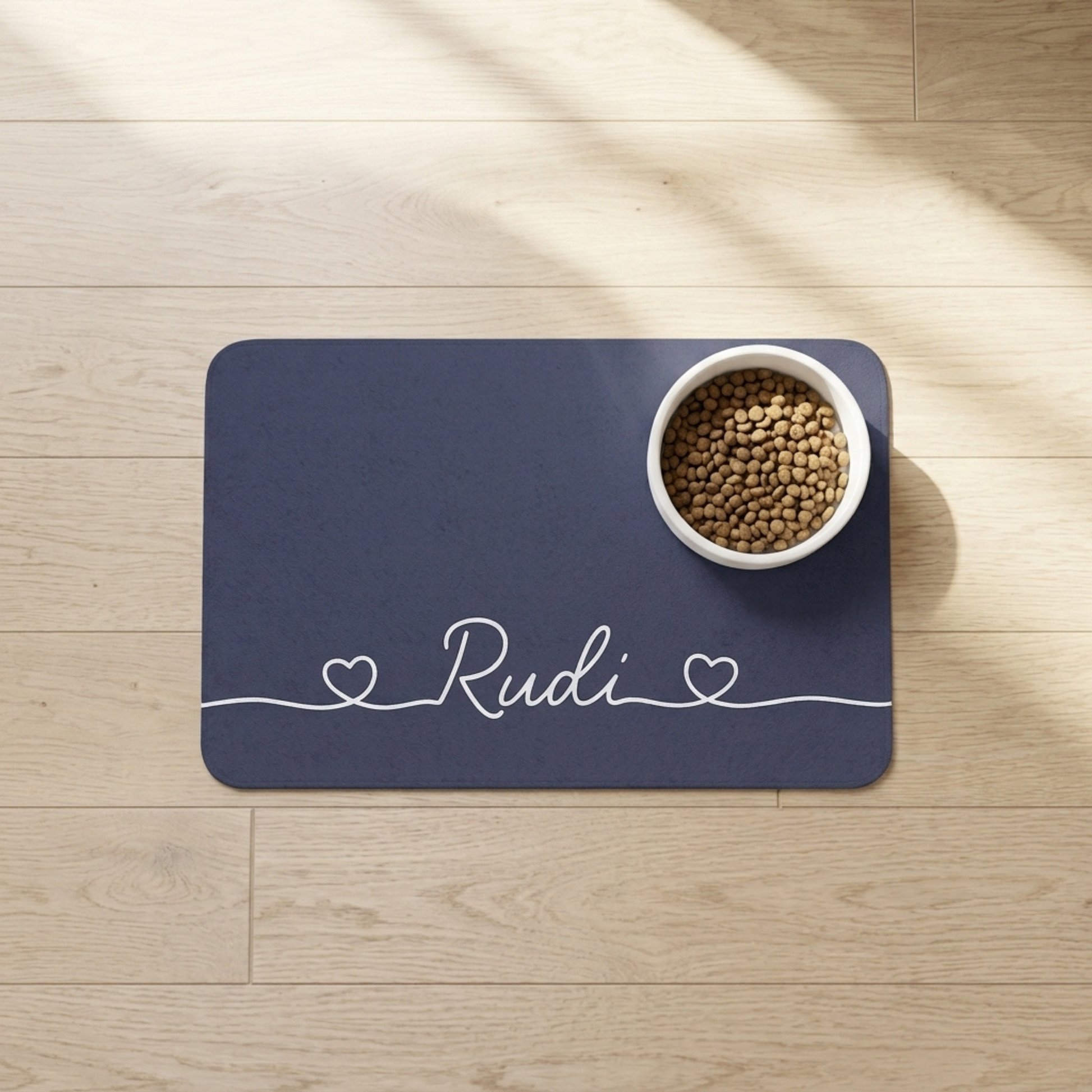 Navy blue dog food mat with 'Rudi' printed on it, featuring a white bowl of pet food on a wooden floor.