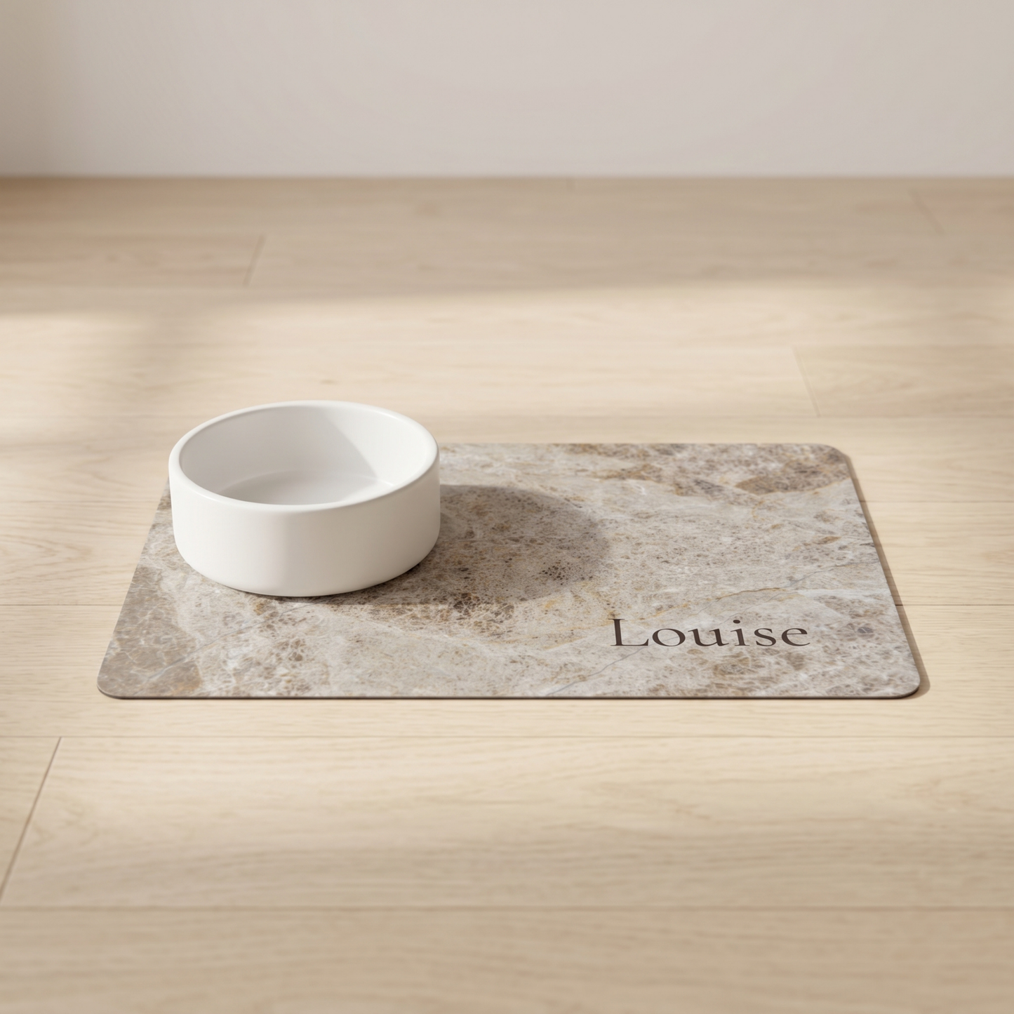 White bowl on a marble-patterned dog food mat with 'Louise' printed on it, placed on a wooden floor.