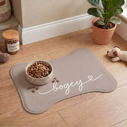 Beige bone-shaped personalized pet feeding mat with a food bowl on a wooden floor next to houseplants