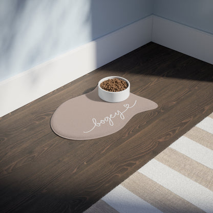 Beige fish-shaped personalized pet feeding mat with a food bowl on a dark wooden floor in sunlight