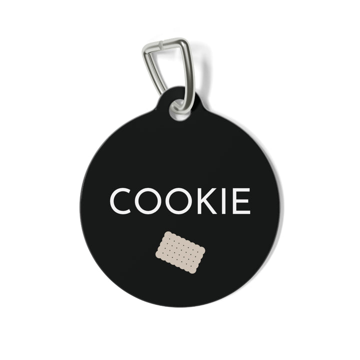 Round black biscuit personalized dog tag with the name Cookie in white capitals and a small sand scalloped biscuit