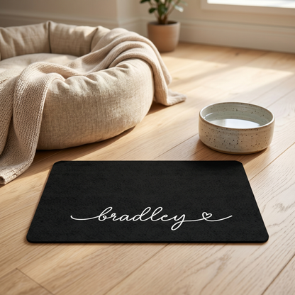 Black personalized pet food mat with a water bowl next to a cozy dog bed on a wooden floor