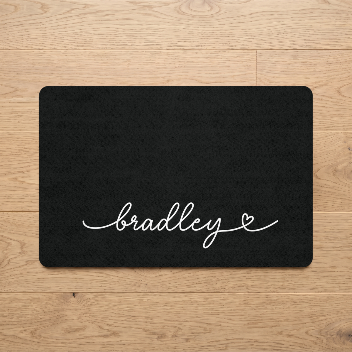 Black personalized pet food mat with the name Bradley on a wooden floor