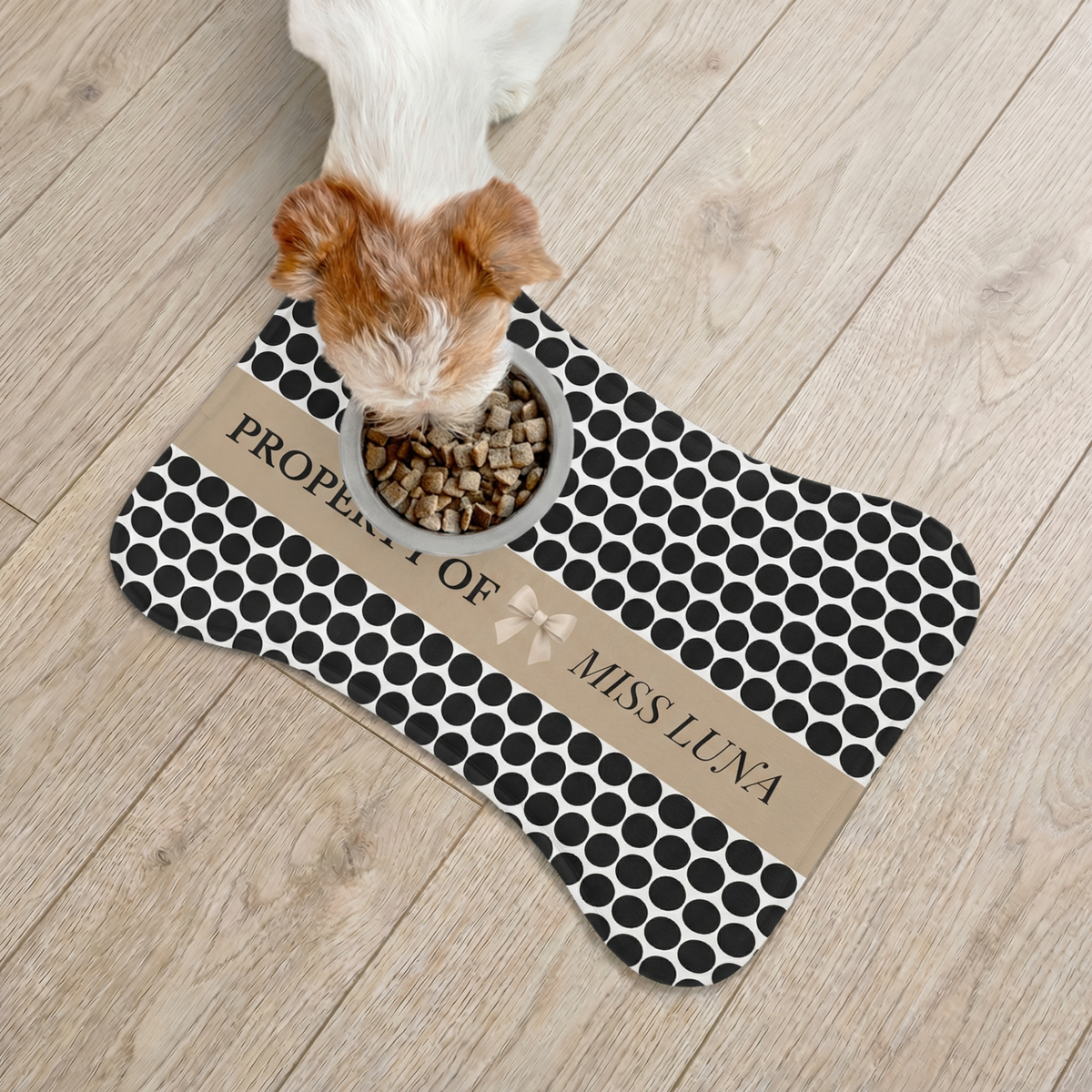 Small dog eating from a bowl on a black polka dot bone-shaped personalized pet feeding mat, seen from above