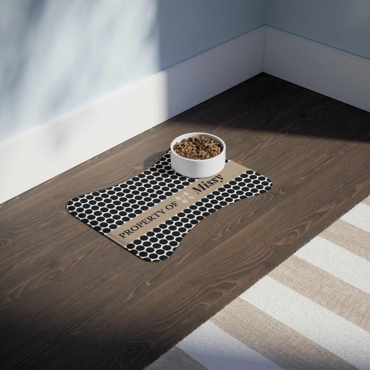 Black polka dot bone-shaped personalized pet feeding mat with a food bowl on a dark wooden floor in sunlight