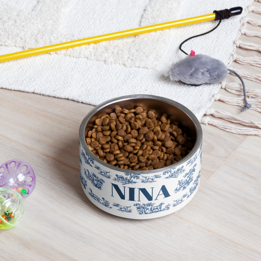 Blue toile personalized dog bowl with the name Nina filled with kibble on a gray floor next to a pet toy