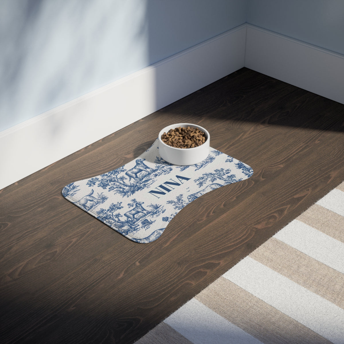 Blue toile personalized pet feeding mat with a food bowl on a dark wooden floor in sunlight