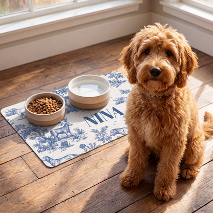 Apricot doodle sitting next to a blue toile personalized pet food mat with food and water bowls