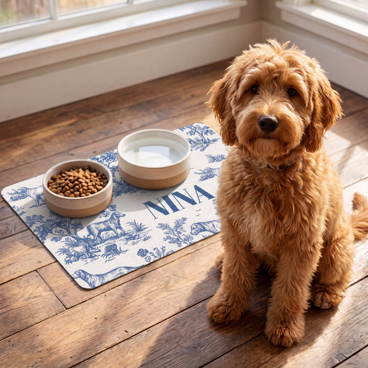Apricot doodle sitting next to a blue toile personalized pet food mat with food and water bowls