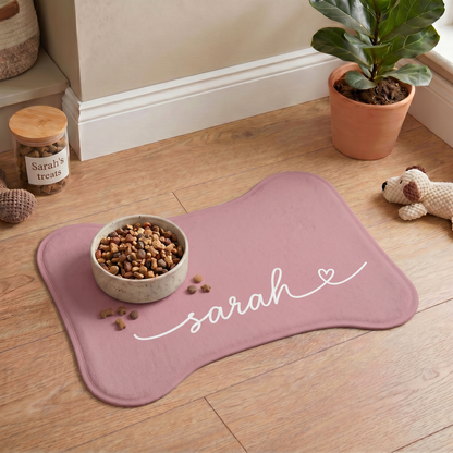 Blush pink bone-shaped personalized pet feeding mat with a food bowl on a wooden floor next to houseplants