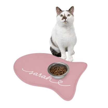 Cat sitting on a blush pink fish-shaped personalized pet feeding mat next to a food bowl