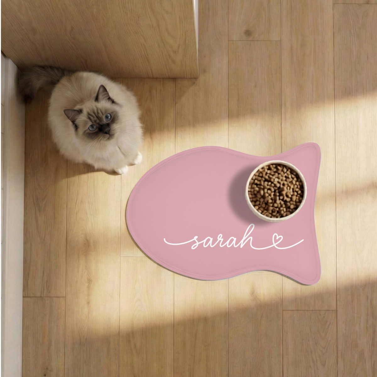Cat next to a blush pink fish-shaped personalized pet feeding mat with a food bowl on a sunlit wooden floor, seen from above