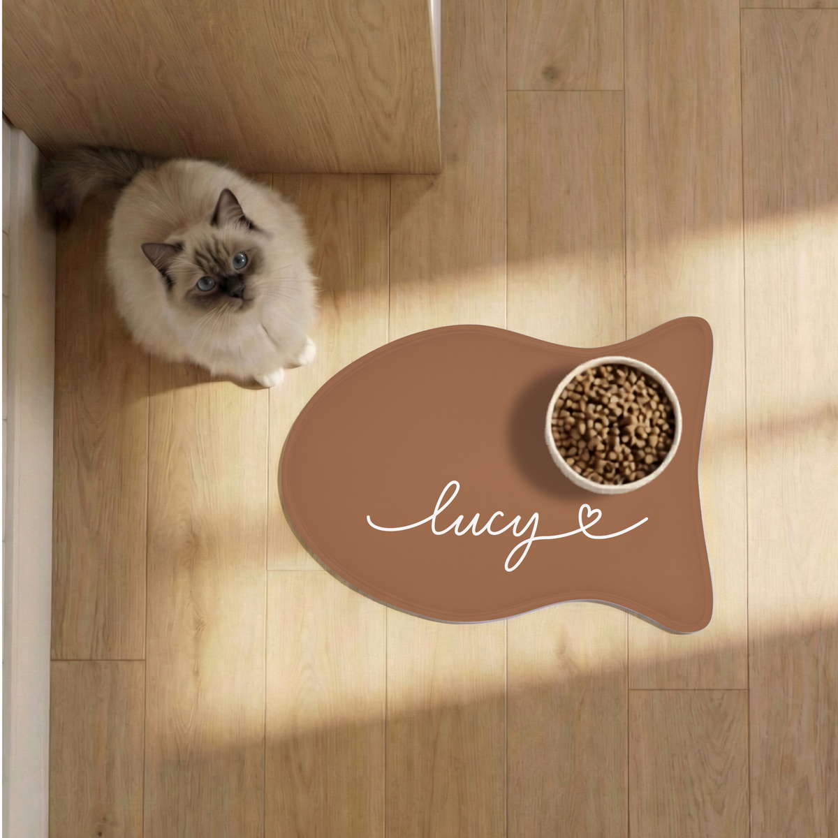 Cat next to a caramel fish-shaped personalized pet feeding mat with a food bowl on a sunlit wooden floor, seen from above