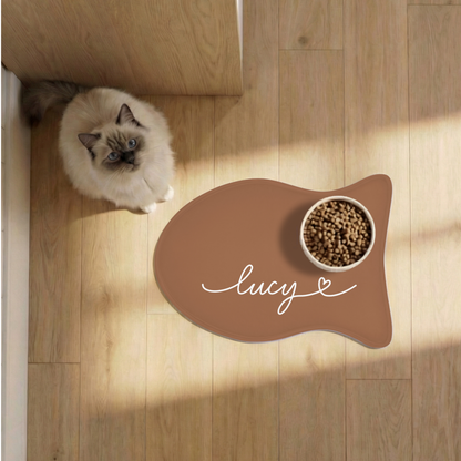 Cat next to a caramel fish-shaped personalized pet feeding mat with a food bowl on a sunlit wooden floor, seen from above