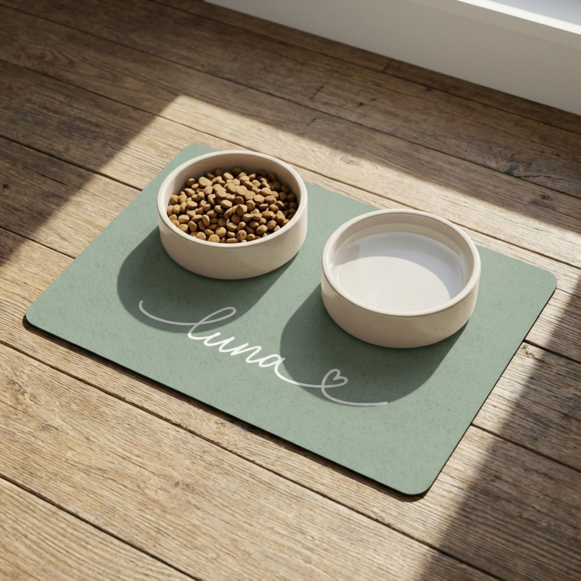 Two bowls on a sage green placemat with 'luna' written on it, one containing dog food on a wooden surface.