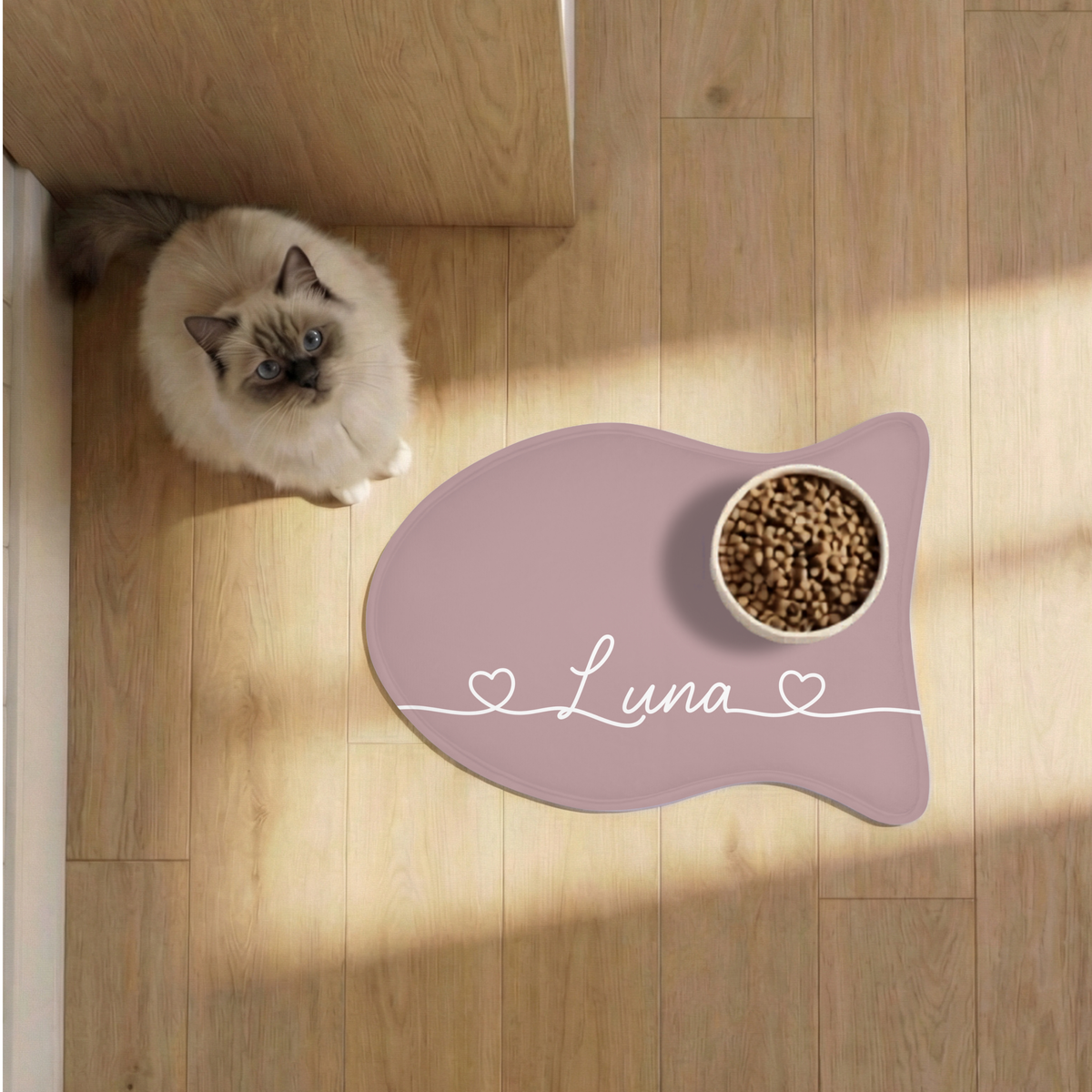 Cat eating from a bowl on a dusty rose fish-shaped personalized feeding mat on a sunny wooden floor