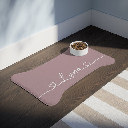 Large dusty rose bone-shaped personalized feeding mat with a bowl on a dark wooden floor in sunlight