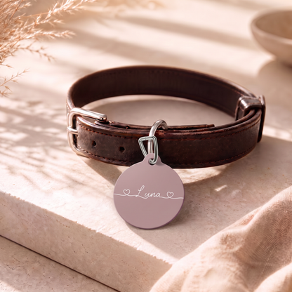 Dusty rose personalized pet tag on a dark leather collar resting on a table