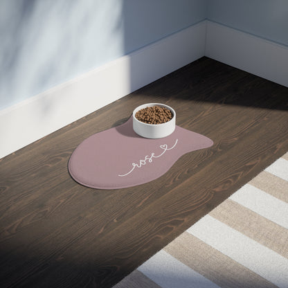 Dusty rose fish-shaped personalized pet feeding mat with a food bowl on a dark wooden floor in sunlight