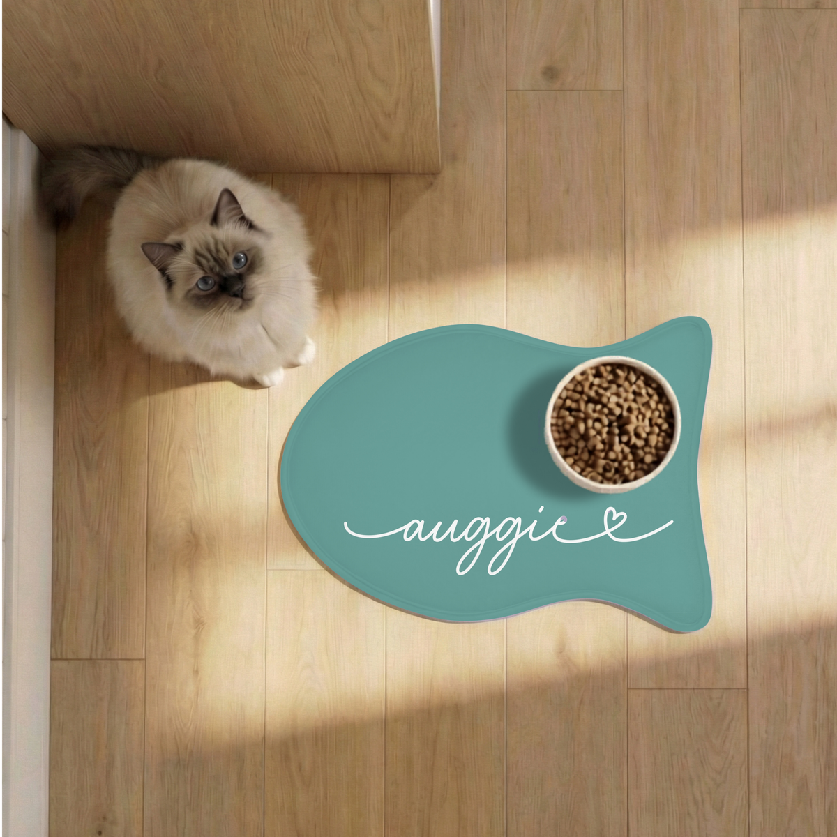 Cat next to a jade fish-shaped personalized pet feeding mat with a food bowl on a sunlit wooden floor, seen from above