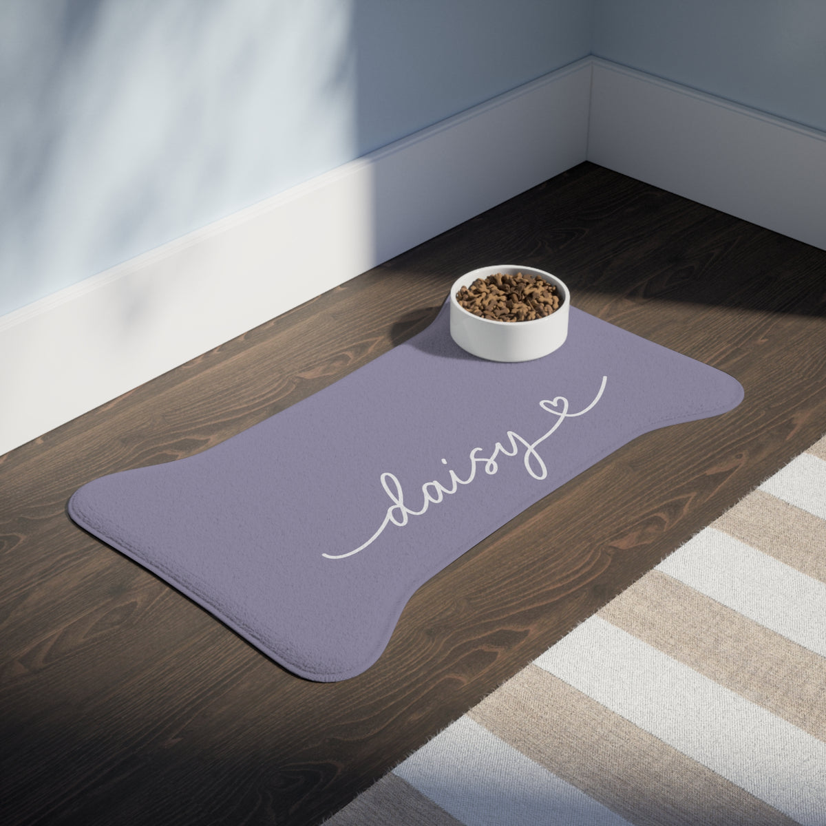 Lavender bone-shaped personalized pet feeding mat with the name Daisy and a food bowl on a dark wooden floor