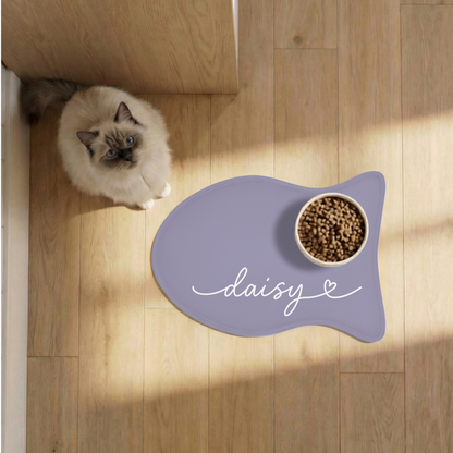 Cat next to a lavender fish-shaped personalized pet feeding mat with a food bowl on a sunlit wooden floor, seen from above