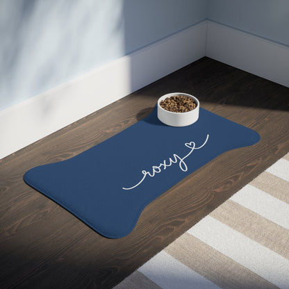 Midnight blue bone-shaped personalized pet feeding mat with the name Roxy and a food bowl on a dark wooden floor