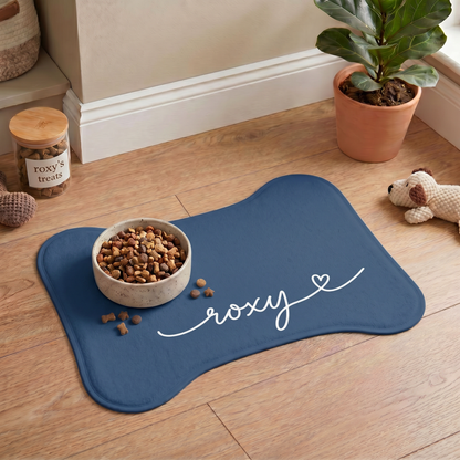 Midnight blue bone-shaped personalized pet feeding mat with a food bowl on a wooden floor next to houseplants