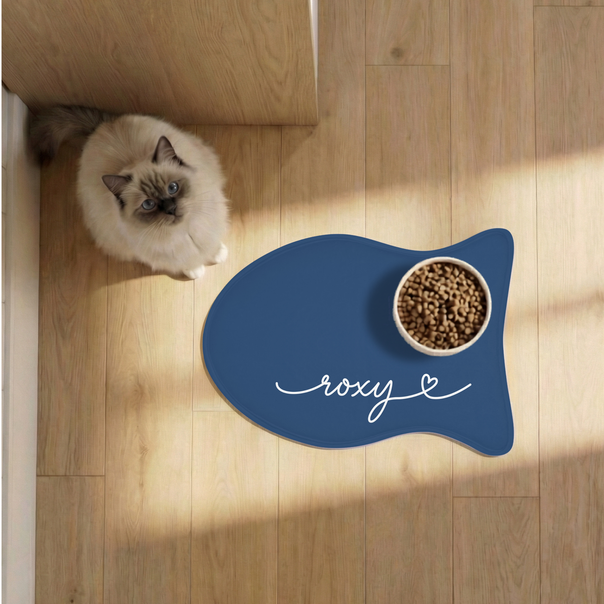 Cat next to a midnight blue fish-shaped personalized pet feeding mat with a food bowl on a sunlit wooden floor, seen from above