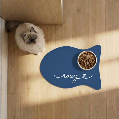 Cat next to a midnight blue fish-shaped personalized pet feeding mat with a food bowl on a sunlit wooden floor, seen from above
