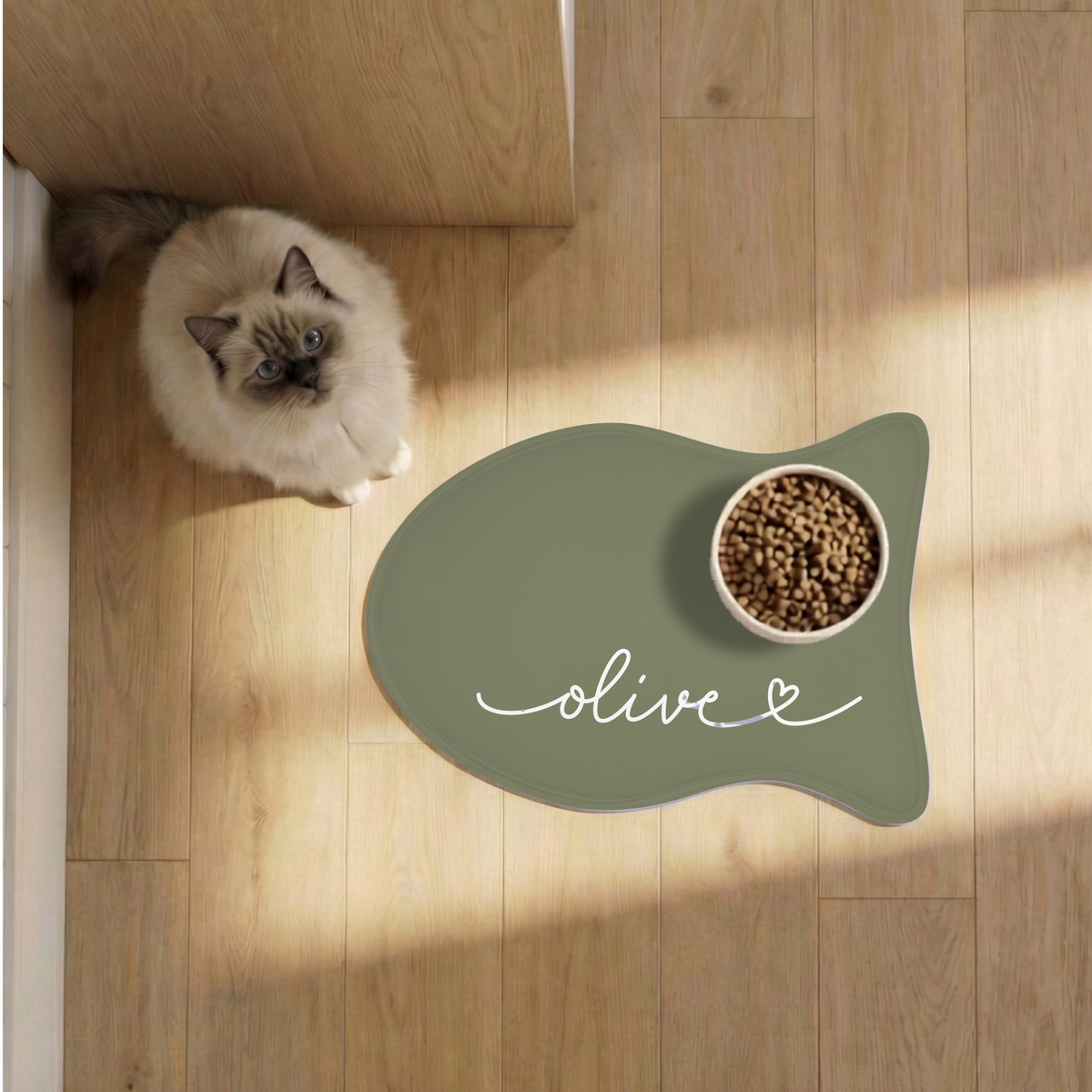 Cat next to an olive green fish-shaped personalized pet feeding mat with a food bowl on a sunlit wooden floor, seen from above