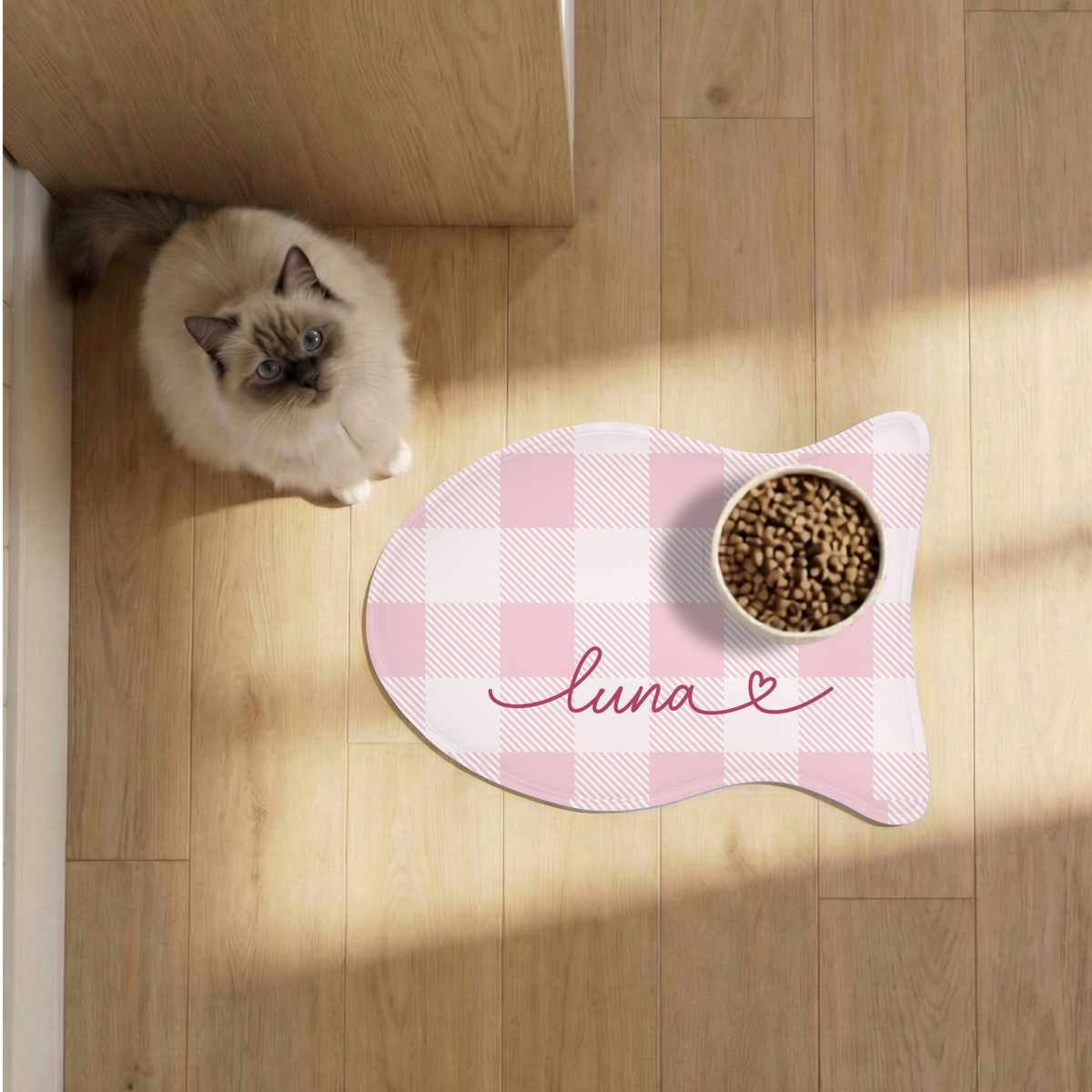 Cat next to a pink checkered fish-shaped personalized pet feeding mat with a food bowl on a sunlit wooden floor, seen from above