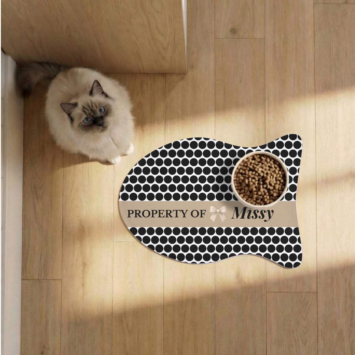 Cat next to a black polka dot fish-shaped personalized pet feeding mat with a food bowl on a sunlit wooden floor, seen from above