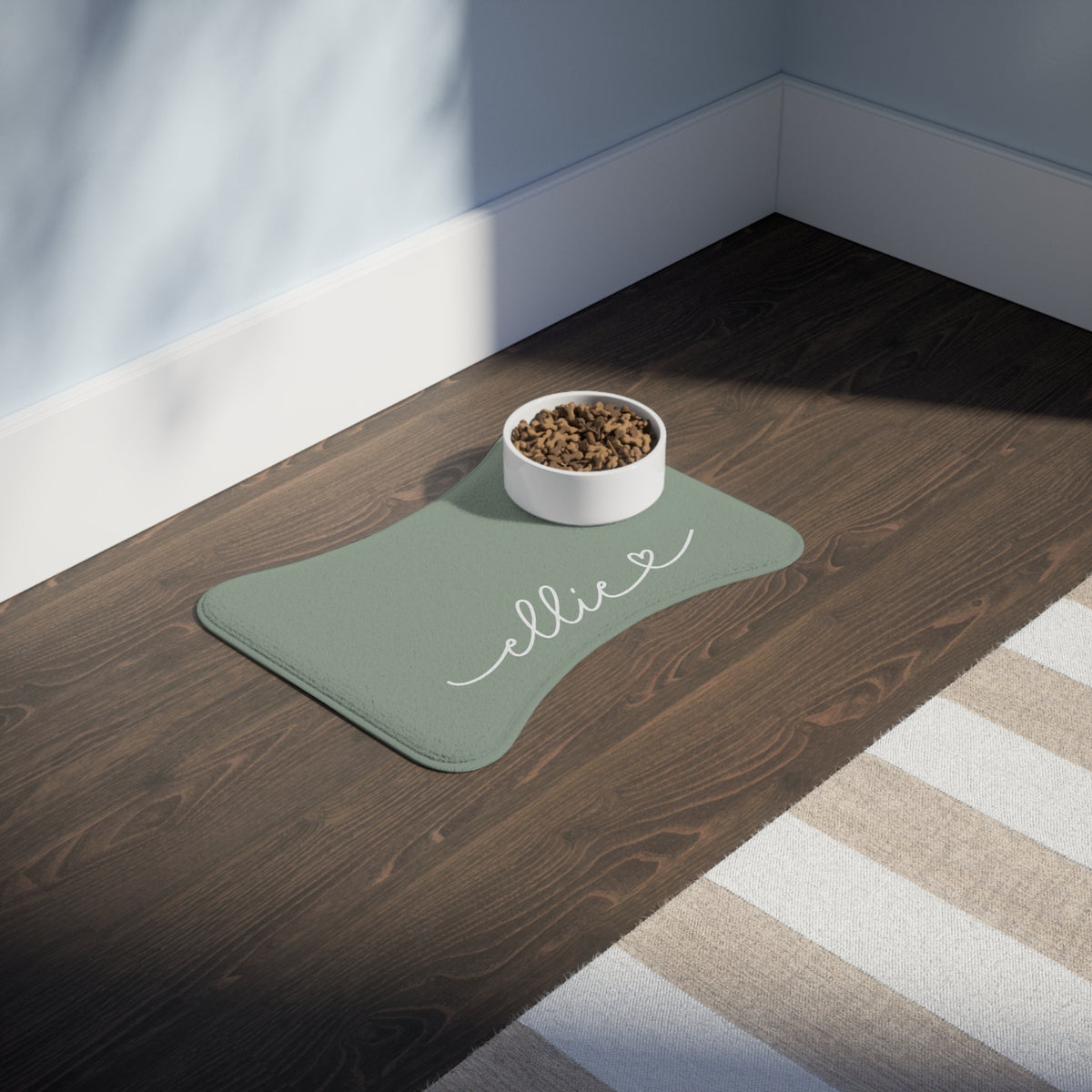 Sage green bone-shaped personalized feeding mat with a food bowl on a dark wooden floor