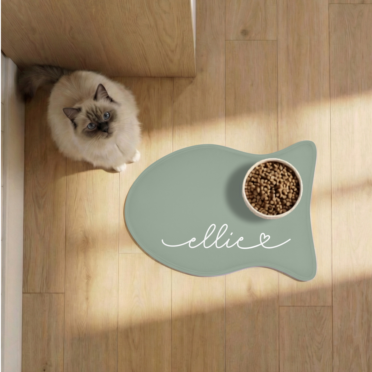 Cat eating from a bowl on a sage green fish-shaped personalized feeding mat on a sunny wooden floor