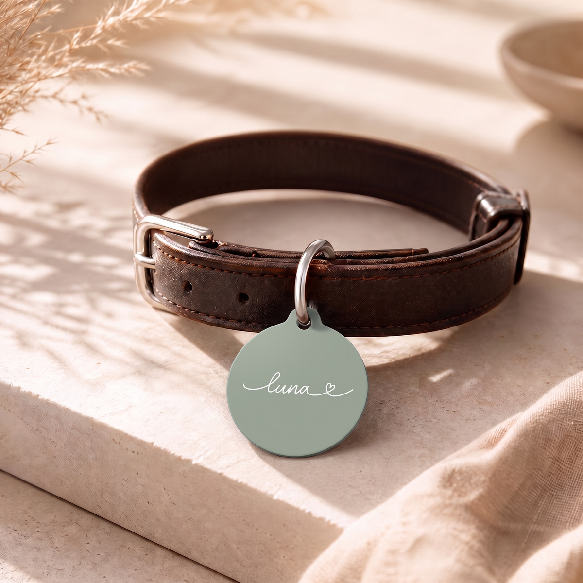 Sage green personalized pet tag on a dark leather collar resting on a table