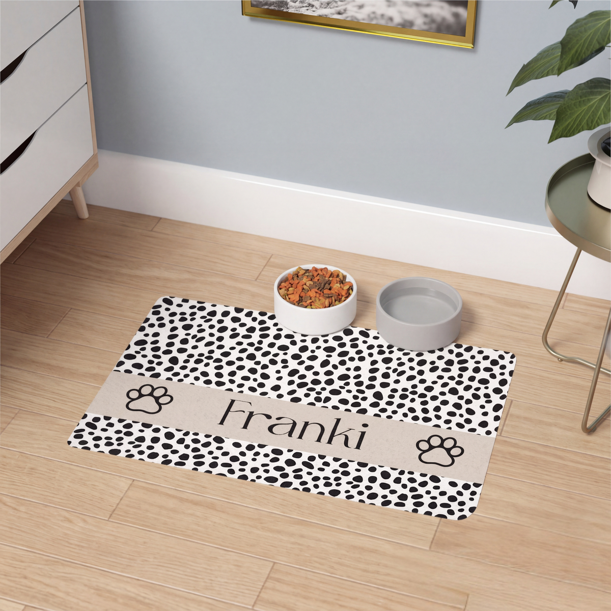 Spotted paw personalized pet food mat with bowls on a wooden floor next to a houseplant