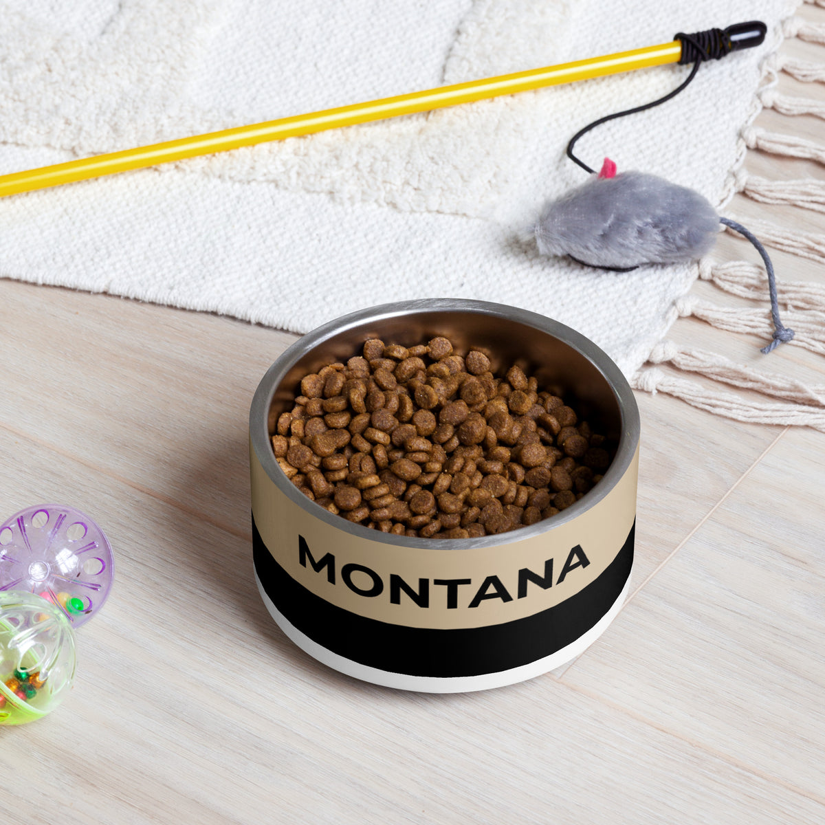 Tan and black two-tone personalized dog bowl filled with kibble on a rug next to a pet toy