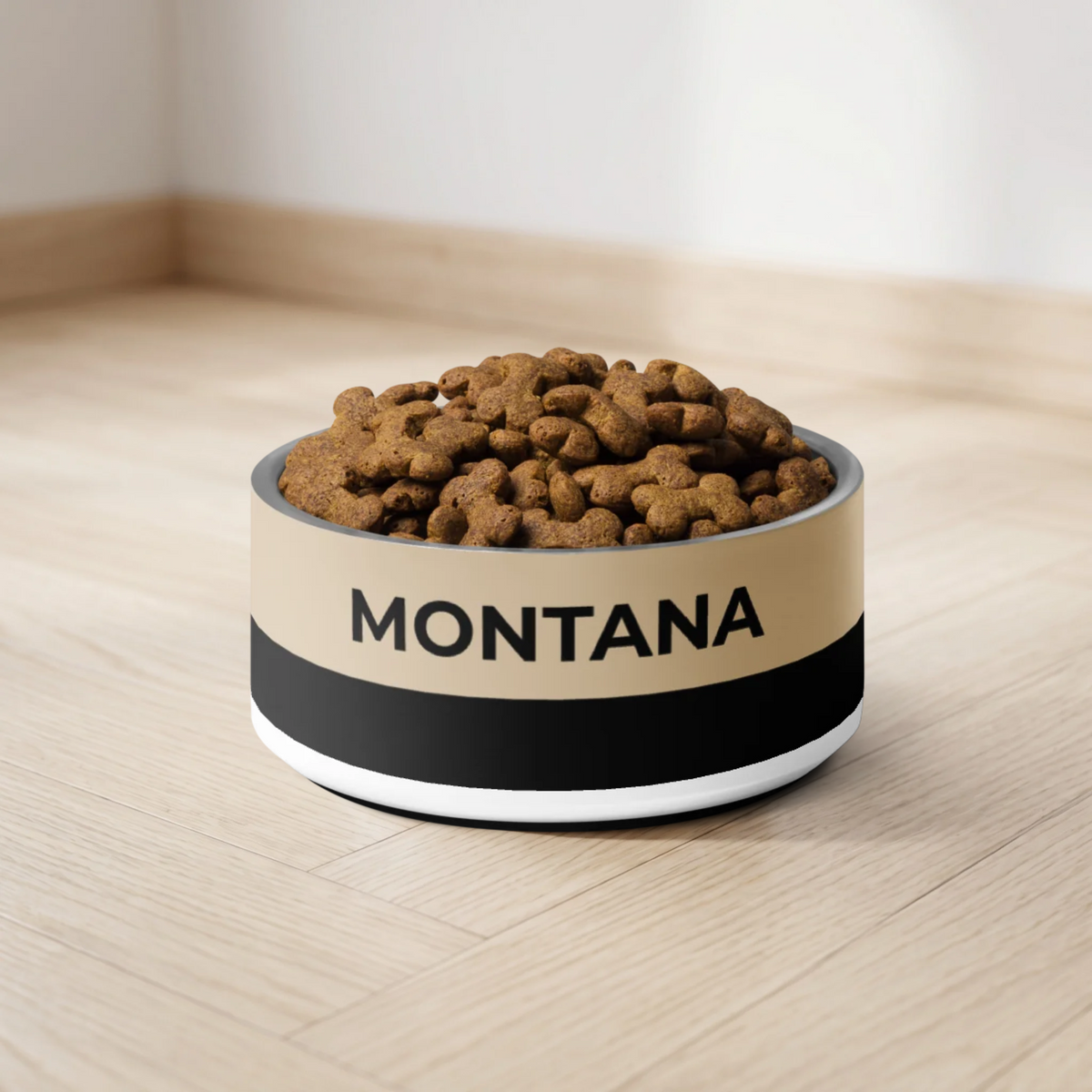 Tan and black two-tone personalized dog bowl with the name Montana filled with kibble on a light wooden floor