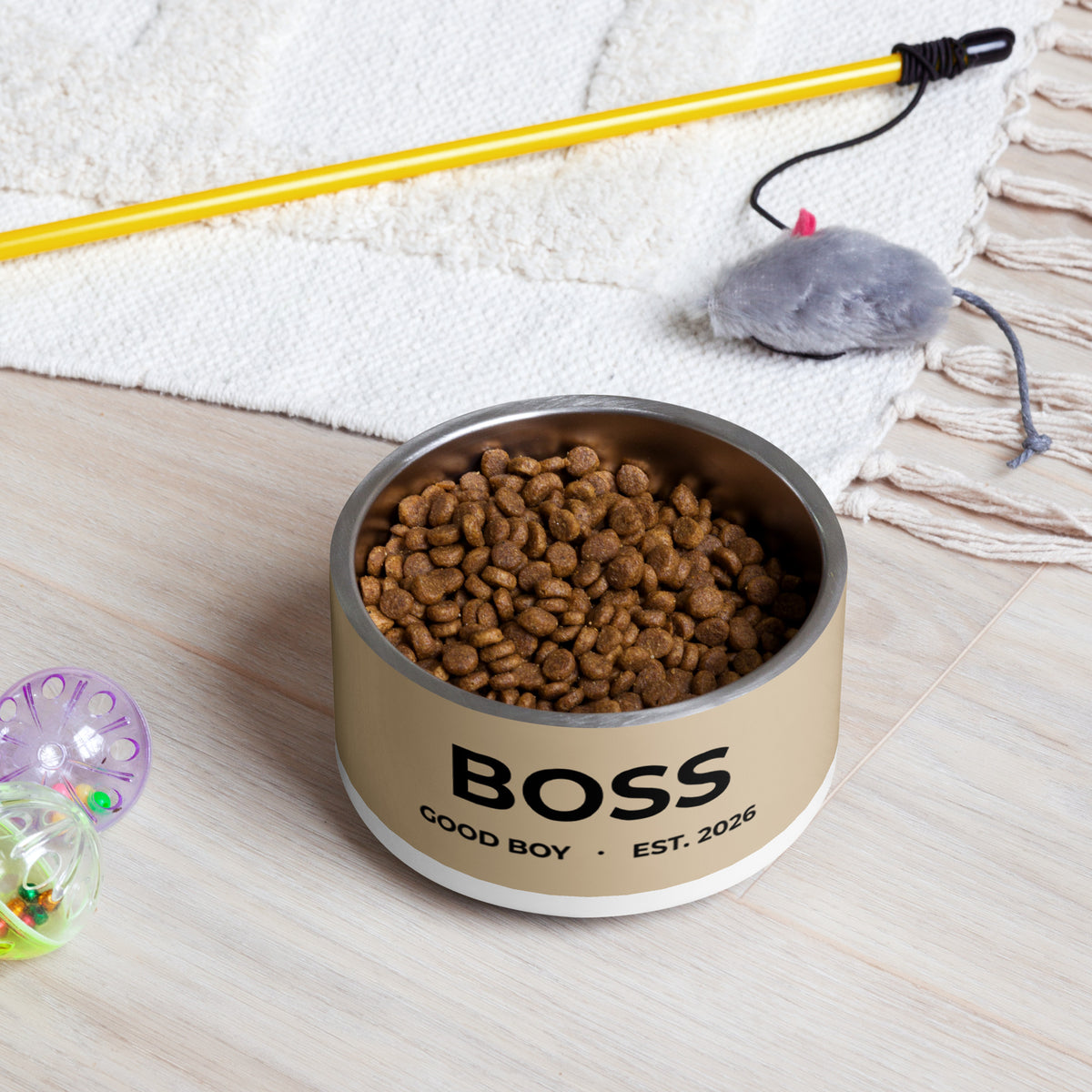 Tan good boy personalized dog bowl filled with kibble on a rug next to a pet toy