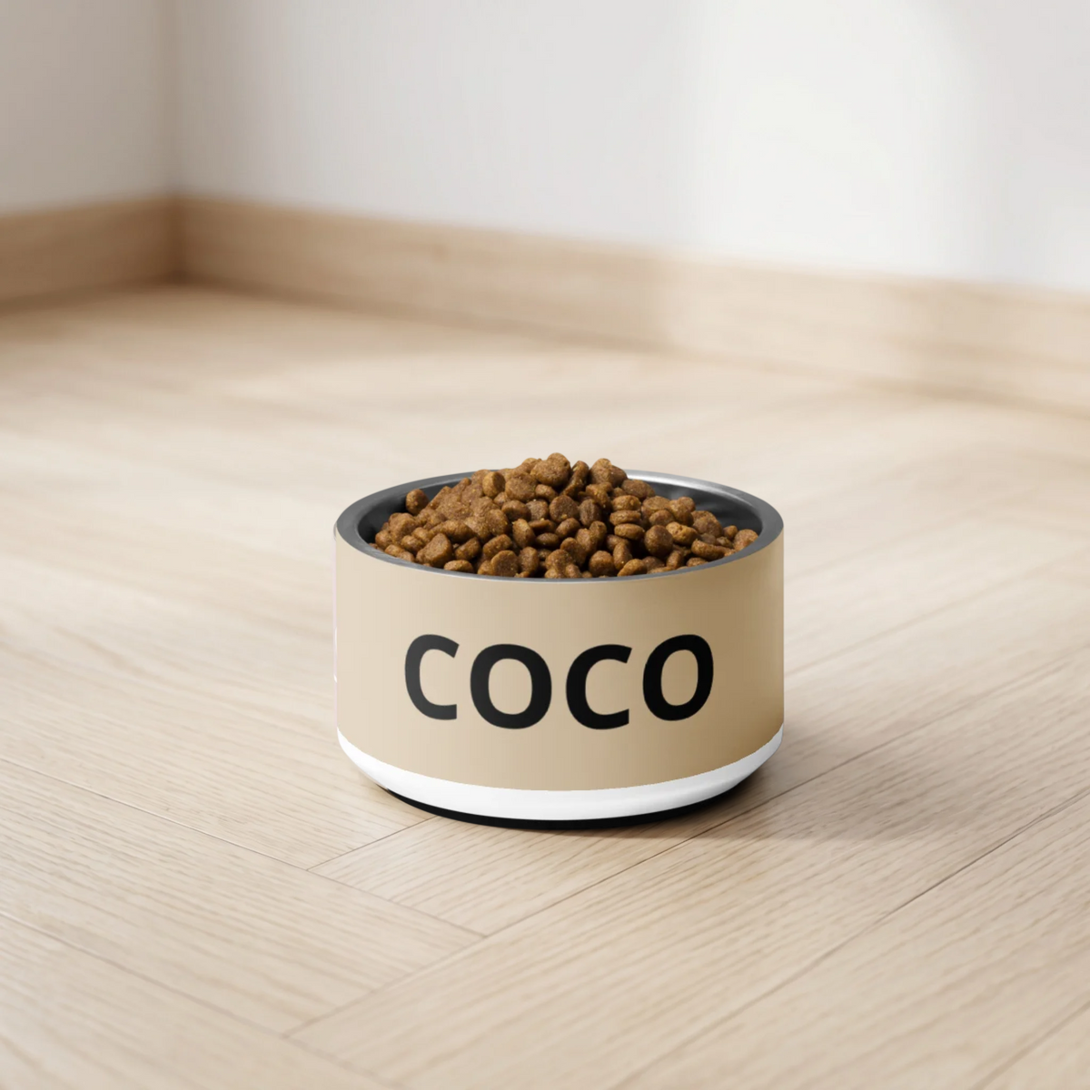 Tan modern personalized dog bowl with the name Coco filled with kibble on a light wooden floor