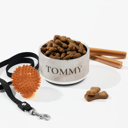 White marble personalized dog bowl with kibble next to a leash, ball and dog treats